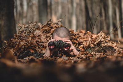 Army soldier looking through binoculars while lying amidst dry leaves in forest