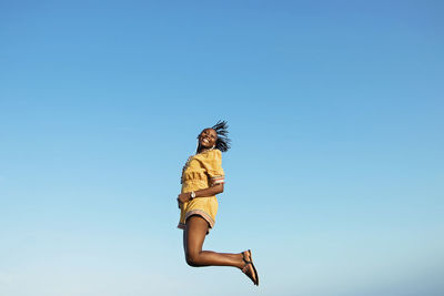 Low angle portrait of cheerful woman jumping against clear blue sky
