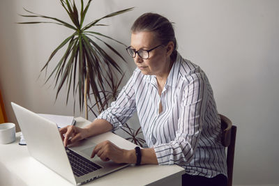 Woman in a white striped shirt aged sits at a computer at home with glasses