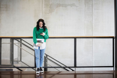 Full length of student using mobile phone while standing at railing against wall in university