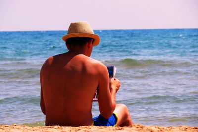 Rear view of shirtless man with magazine while relaxing on shore at beach