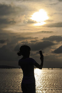 Woman photographing through mobile phone while standing against sea during sunset