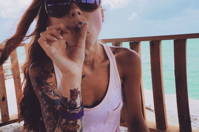 Portrait of young woman wearing sunglasses smoking against sea