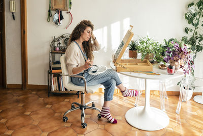 Woman with hand in hair sitting on chair painting in living room at home