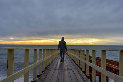 Rear view of man standing on railing against sea during sunset