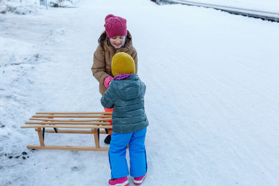Elder sister helping yonger sister on snow slope with wooden sleigh
