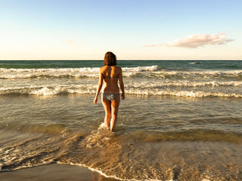 Rear view of woman wearing bikini walking in sea against sky during sunset