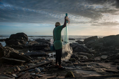 Full length of fisherman with net at rocky coastline against sky during sunset