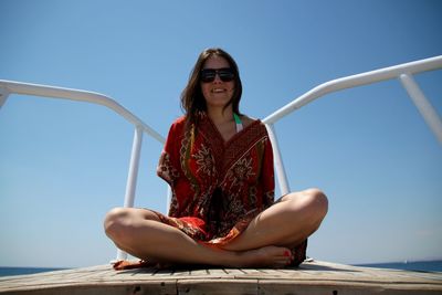 Full length of young woman sitting on ship bow against clear blue sky at sea