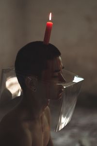 Shirtless man with lit candle on head and covered face in room