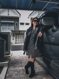 A girl wearing a polkadot dress, jacket, sun glasses and high boots standing outside the car 