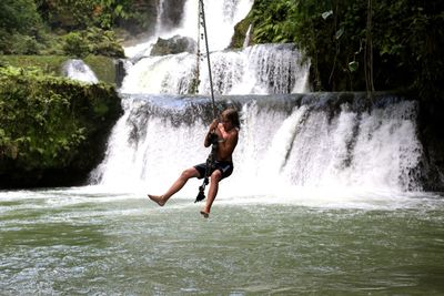 Boy swinging on rope against waterfall