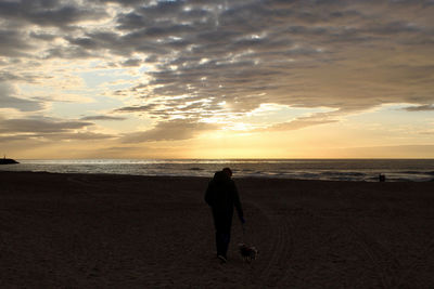 Rear view of silhouette standing on beach against sky during sunset