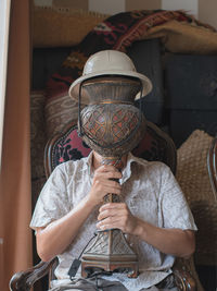 A man hides his face with a vintage red table lamp base under a beige hat