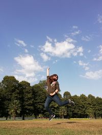 Happy mature woman with arms raised jumping on field against sky