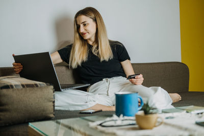 Smiling woman using laptop while holding credit card on sofa