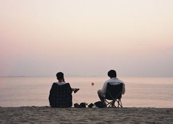 Rear view of men sitting on beach against sky during sunset