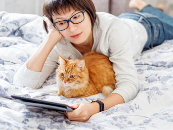 Cute ginger cat and woman in glasses are lying in bed with tablet. 