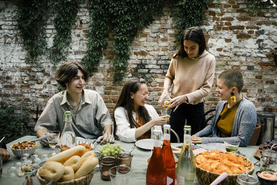 Woman pouring drink to female friend sitting at dining table during garden party in back yard