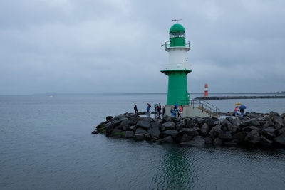 People standing below lighthouse by sea against sky