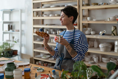 Artisan tattooed girl drink coffee and looking at tasty bun lustily in art studio during break