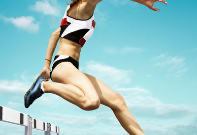 Low section of woman jumping over hurdle against blue sky