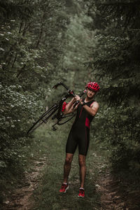 Portrait of young man carrying bicycle while standing on field amidst trees in forest
