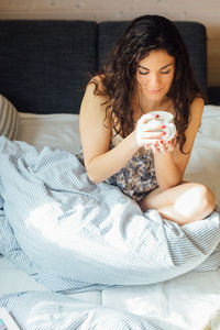 High angle view of young woman having coffee while sitting on bed at home