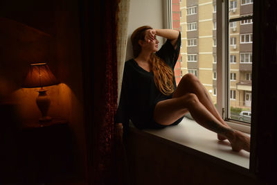 Young woman looking away while sitting on window at home