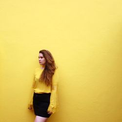 Young woman looking away while standing against yellow wall