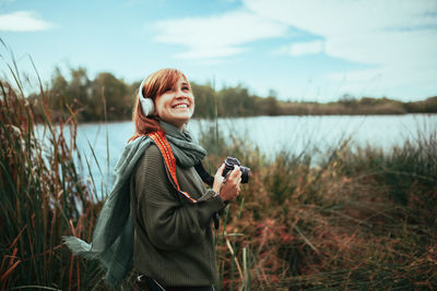 Smiling young woman holding camera standing on grassy land