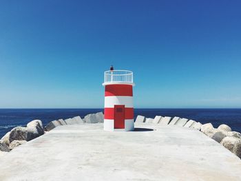 Red and white striped lighthouse by sea against clear blue sky