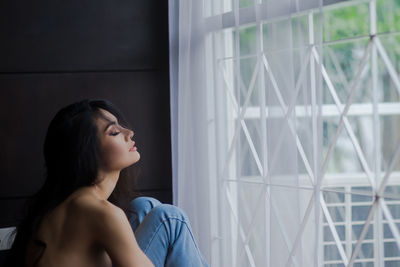 Side view of shirtless young woman with eyes closed sitting by window at home