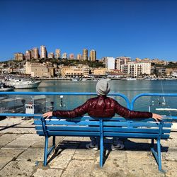 Empty bench by river in city against clear blue sky