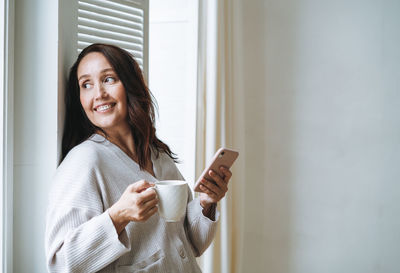 Young woman in cozy cardigan with cup of tea in hands using mobile phone in bright interior at home