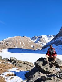 Man sitting on rock against snowcapped mountains against clear blue sky