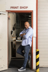 Smiling employee with arms crossed looking down while leaning on doorway at print shop