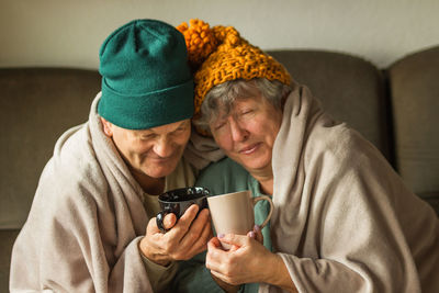 Grandparents in knitted caps wrapped in warm blanket hold cups of hot tea, feeling cold at home.