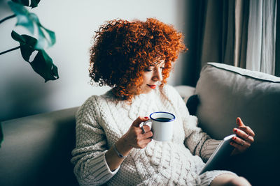 Young woman holding coffee while sitting on sofa at home