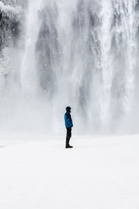 Side view of man standing on snow covered field against waterfall