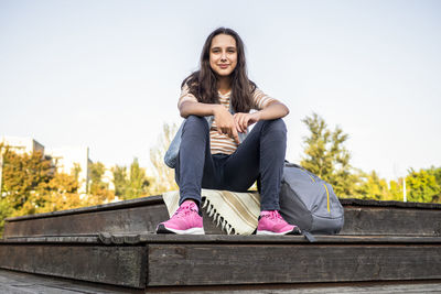 Portrait of young woman sitting on bench against clear sky