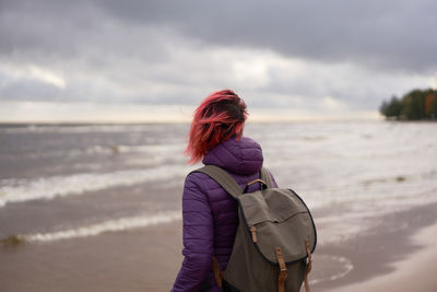 Back view of unrecognizable female traveler in outerwear standing on shore and admiring calm sea on cloudy day in sestroretsk in autumn