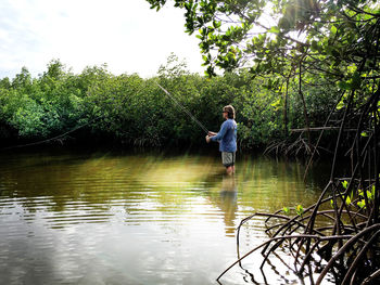 Man fishing at river by trees against sky