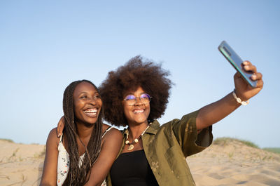 Cheerful young african american female friends smiling brightly while taking selfie on smartphone during summer weekend on sandy seashore