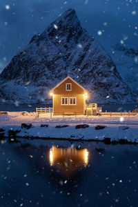 Illuminated house by lake against snowcapped mountain