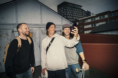 Smiling male friends taking selfie on smart phone against shed on street