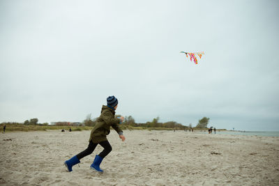 Rear view of boy holding kite running on beach against sky