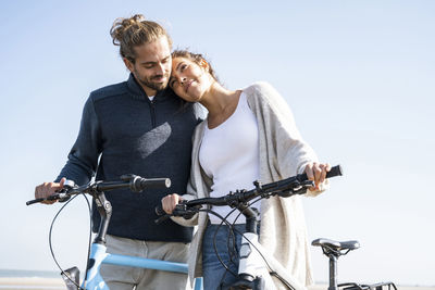 Beautiful woman with head on boyfriend's shoulder standing with bicycles at beach against clear sky on sunny day
