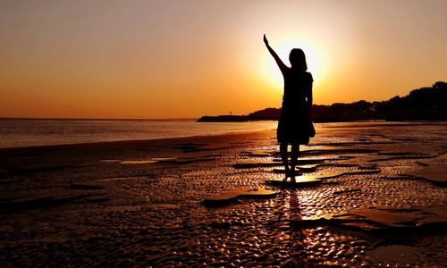 Silhouette woman with arms raised standing on beach against sky during sunset