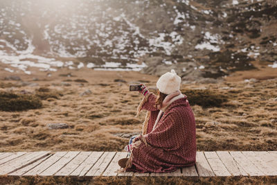 Side view of woman using mobile phone while sitting on footpath against mountain and sky